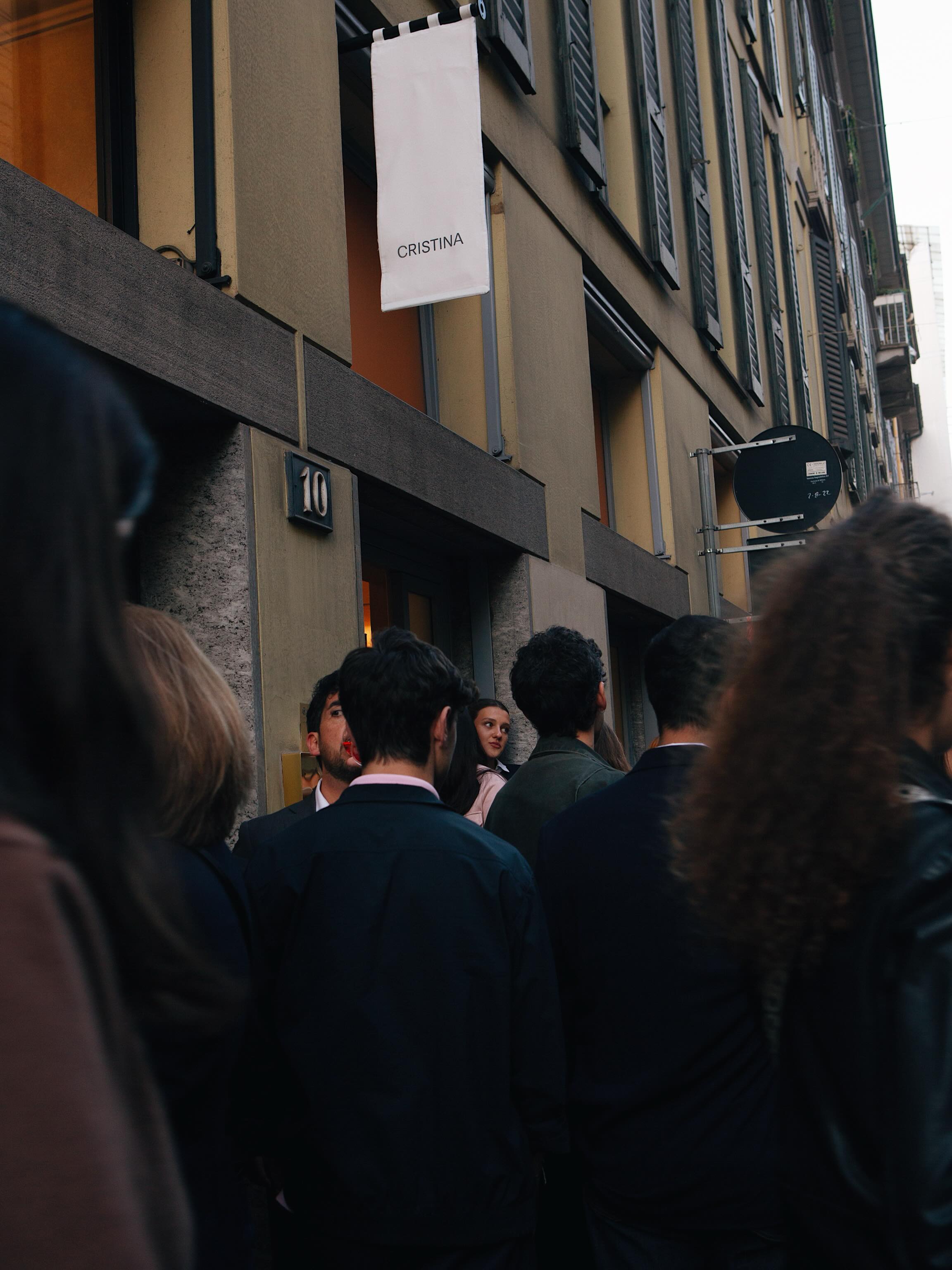 A large crowd outside the CRISTINA showroom underneath a white flag with the CRISTINA logo